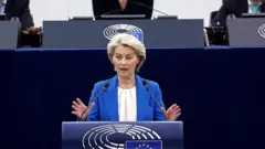 Mavi takım elbiseli sarışın bir kadın AB bayrağı olan bir kürsüde konuşuyor.