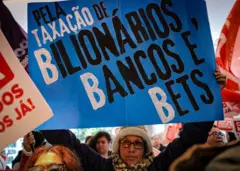 Manifestante durante protesto na sede do banco Itaú BBA, em São Paulo, com um cartaz onde se lê: "Pela taxação de bilionários, bancos e bets"