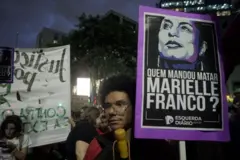 Em manifestação à noite, com outras pessoas ao fundo, jovem segura cartaz com fotosinais greenbetsMarielle e pergunta: 'Quem mandou matar Marielle Franco?'