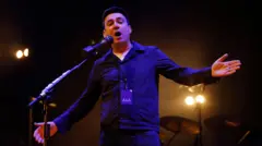 Andy Burnham no palco, com os braços abertos. Uma bateria aparece ao fundo, com luzes de palco iluminando-o. Ele está vestindo uma camiseta branca e uma camisa roxa de manga comprida com botões, em 13 de março de 2023.