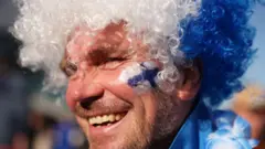 Homem sorridente ao ar livre, usando uma peruca cacheada nas cores branco e azul e com o rosto pintado com tinta azul e branca na bochecha. Ele está em um campeonato de tênis, com a luz solar direta iluminando seu rosto e outras pessoas desfocadas ao fundo.
