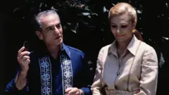 Homem está usando uma camisa azul e tem cabelos grisalhos. Uma mulher está ao lado dele usando uma camisa clara e cabelos presos.