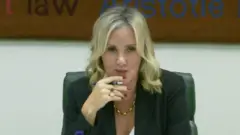 Caroline Dinenage - cabelo loiro, unhas vermelhas, segurando uma caneta, jaqueta escura, corrente dourada, sentada em uma cadeira.