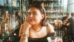 バーにいる若い女性の上半身の写真。黒いトップスを着て、あごを手に乗せている。サングラスで前髪をあげていて、金色のイヤリング、ネックレス、ブレスレットを身につけている。背後にある棚には飲み物のボトルがたくさん並んでいる