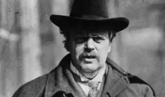 Fotografia em preto e branco do filósofo G K Chesterton, um homem branco de bigode usando uma cartola preta e óculos 