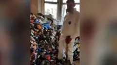 La fotografía muestra una pila de basura apilada en el ventanal. Un joven está de pie frente a la pila. Lleva equipo de protección personal completo, mascarilla y una pala en la mano. Lleva guantes negros y rojos.