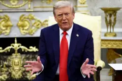 ترامب محركاً يديه ويتحدث. يرتدي بدلة رسمية كحلية اللون وقميصاً أبيض وربطة عنق حمراء، يجلس على كرسي يميل إلى اللون الذهبي في البيت الأبيض.