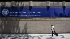 Um homem de camisa branca caminha pela calçada em frente a um muro com uma placa que diz “Banco Central da Venezuela”