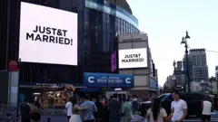 Pessoas assistem a telões gigantes exibindo a mensagem "JUST&T MARRIED!" (T&T de Taylor e Travis) do lado de fora do Madison Square Garden, local onde supostamente aconteceria a cerimônia de casamento da cantora pop Taylor Swift e do atleta profissional Travis Kelce, na cidade de Nova York, em 3 de julho de 2026.