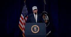 US President Donald Trump speaks at a podium with the US Seal while waring a dress shirt, dark blazer and white hat that is emblazoned with USA.