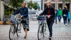 Dos mujeres caminan sonrientes empujando sendas bicicletas por una calle peatonal de Oslo. 