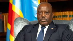 Joseph Kabila wahoze ari perezida wa DR Congo yicaye mu ntebe yambaye isuti y'umukara inyuma ya hari ibendera rya RD Congo