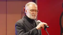 Sergei Mikheev veste terno cinza e gola alta preta. De óculos, com cabelos grisalhos curtos e barba rente ao rosto, ele aparece em um palco segurando um microfone