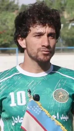 Faryob viloyatining "Sari sabz" futbol jamoasi futbolchisi Nozir Ahmad Nazariy makeraga qarab, qo'lida O'zbekiston bayrog'ini ushlab turibdi, Sochlari to'q jigar rang, egnida furtbol maxsus kiyimi. 