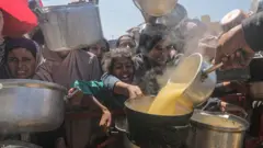 Palestinos desalojados internamente, incluindo crianças, seguram panelas enquanto recebem comida de uma cozinha beneficente, na Cidade de Gaza, na Faixa de Gaza, em 4 de agosto de 2025