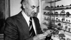 O cientista suíço Albert Hofmann, de terno e gravata, examinando pedras em um mostruário.