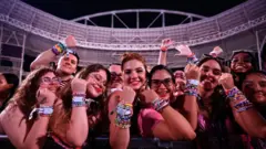 A imagem mostra um grupo de fãs de Taylor Swift posando na grade de um estádio antes de um show, exibindo dezenas de pulseiras coloridas nos pulsos. Sorrindo para a câmera, eles levantam os braços para destacar os acessórios, que se tornaram uma marca registrada entre admiradores da cantora, tendo ao fundo a estrutura iluminada da arena onde acontecerá a apresentação.