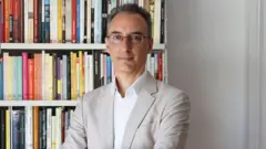 O jornalista e escritor Andrea Rizzi, com terno claro e óculos, em frente a uma estante com livros