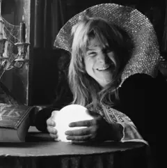 Ozzy Osbourne segurando uma bola de cristal vestido como um mago. Na mesa à sua frente, há um livro de feitiços e uma vela com teias de aranhas
