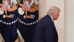 Trump caminha para a direita e se afasta da câmera. À esquerda, há duas bandeiras dos EUA