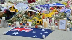 محل یادبود قربانیان حمله در ساحل بوندی استرالیا. پرچم استرالیا در جلو تصویر و دسته‌های گل و شمع در عقب دیده می‌شوند. 