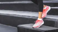 A woman wearing black leggings and pink athletic shoes, standing on a set of black steps. One foot is placed on a higher step while the other remains on a lower one, suggesting an exercise or stretching.
