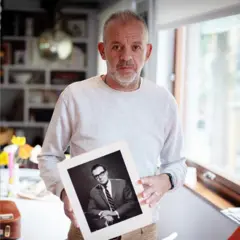 Antony Easton, vestindo uma camisa de manga comprida de cor clara, está em pé dentro de casa, segurando uma fotografia em preto e branco de seu pai, Peter Easton, vestido com terno e gravata. Atrás dele, há uma sala com estantes de livros e vários objetos, iluminada pela luz natural que entra por uma janela à direita.