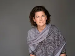 Mulher de cabelo curto e levemente ondulado, vestindo um xale cinza com estampa floral por cima, posando diante de um fundo neutro