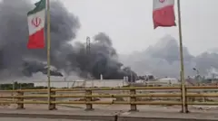 Дым поднимается в воздух после ударов по нефтехимической зоне Махшахр в округе Бандар-Махшахр провинции Хузестан, Иран. На переднем плане у дороги на флагштоках развеваются два иранских флага, а позади них виден подвергшийся удару завод