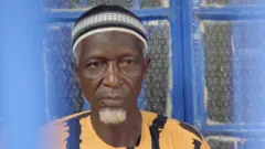 Foday Musa, who has a white goatee, looks solemn. He wears a yellow and black top and white and black knitted skull cap and behind him is a window with blue painted frames.