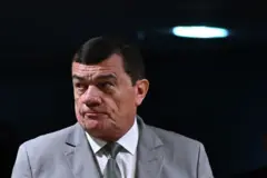 Nogueira olhando para o lado, com olhar tenso e em pé; atrás, um fundo preto