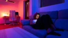 Jovem recostada no sofá olhando para uma tela, com as luzes da sala reduzidas e uma mesa com abajur aceso ao fundo