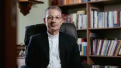 Padre Evandro Campos, reitor do seminário de Belo Horizonte, sentado numa cadeira de escritório com livros ao fundo