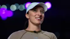 Polska tenisistka Iga Świątek na czarnym tle. Uśmiecha się, a na głowie ma białą czapkę z daszkiem.