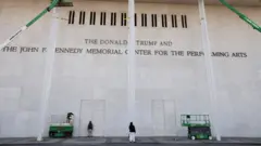 ベージュ色の石の壁に、緑色のフォークリフトに乗った作業員が、「The John F. Kennedy Center for the Performing Arts」という文字の上に「The Donald Trump and」という文字を足した後の様子