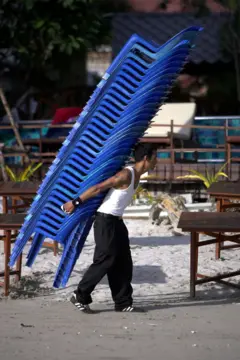Homem carrega uma pilha de cadeiras monobloco de cor azul.