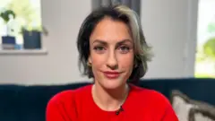 Madelaine Thomas sits on a dark blue couch and smiles at the camera. She is wearing a red short-sleeved dress. He has short, dark hair with a blonde streak in the front.