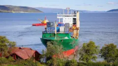 Uma vista ampla mostrando a casa de telhado vermelho, que parece pequena perto do navio e nem chega ao convés. A casa é cercada por árvores e o céu ao fundo é azul.