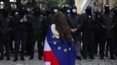 A young woman draped in Georgian and EU flags stands in front of a line of riot police