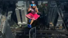 A girl looks through an eclipse viewer on an elevated, transparent platform, with buildings and roads in the distance below her