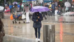 Moça de máscara e casaco de frio caminha sob a chuva com guarda-chuva, ao gfundo, diversas pessoas na rua