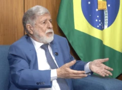 Foto de Celso Amorim gesticulando com as mãos e com uma bandeira do Brasil do lado direito da tela