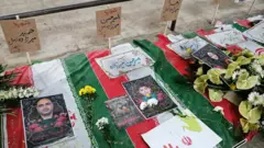 Uma fileira de bandeiras cobre sepulturas com fotos e flores sobre elas