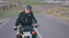 یوه ښځه په یوه تش سړک موټرسايکل ځغلوي او شاته یې شنې غونډۍ ښکاري.