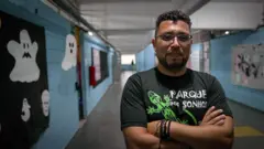 Régis Marques, con gafas y una camiseta con un diseño, mira a cámara con los brazos cruzados. Está en un pasillo de la escuela que dirige desde 2016.