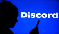 A sombra de uma pessoa de perfil usando o celular, em frente a um fundo azul com o logotipo do Discord