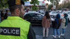 یک پلیس در خیابانی در تهران به اطرافش نگاه می‌کند در حالی که چند دختر بی‌حجاب در رفت و آمدند