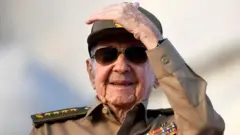 カーキ色の軍服姿でサングラスをかけたカストロ氏が左手を上げほほ笑んでいる