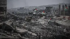 Pilhas de escombros na Faixa de Gaza, sobre as quais se veem bandeiras palestinas. Ao fundo, o terreno está totalmente destruído e coberto de escombros entre ruínas de edifícios