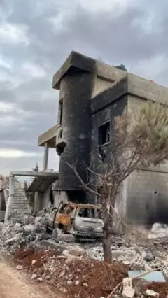 Un edificio de piedra de tres plantas, ennegrecido y derruido, la casa familiar de Qassem, en Líbano, tras los bombardeos, yace en ruinas, con los restos calcinados de un coche y un árbol entre los escombros del exterior, bajo un cielo nublado.